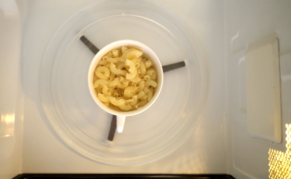 A small white cup filled with uncooked macaroni sits centered in the microwave