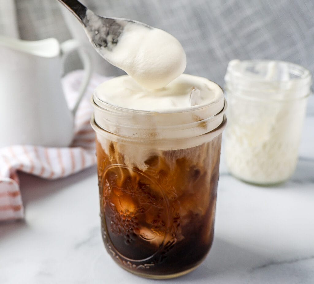 A glass of iced coffee topped with sweet cream cold foam being spooned on, with a jar of foam and a pitcher in the background