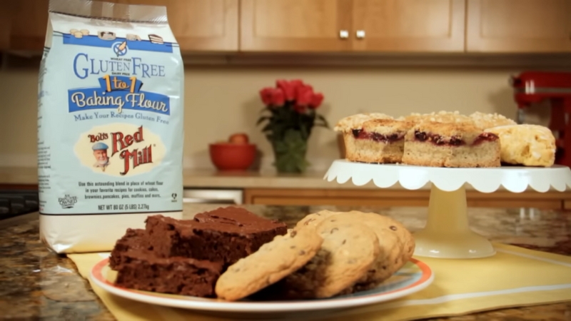 A Package of Bob’s Red Mill Gluten Free 1-To-1 Baking Flour with Freshly Baked Brownies, Cookies, and A Plate of Scones in The Background