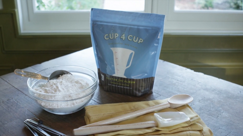 A Package of Cup4Cup Multipurpose Gluten-Free Flour Next to A Bowl of Flour and Wooden Measuring Spoons on A Countertop