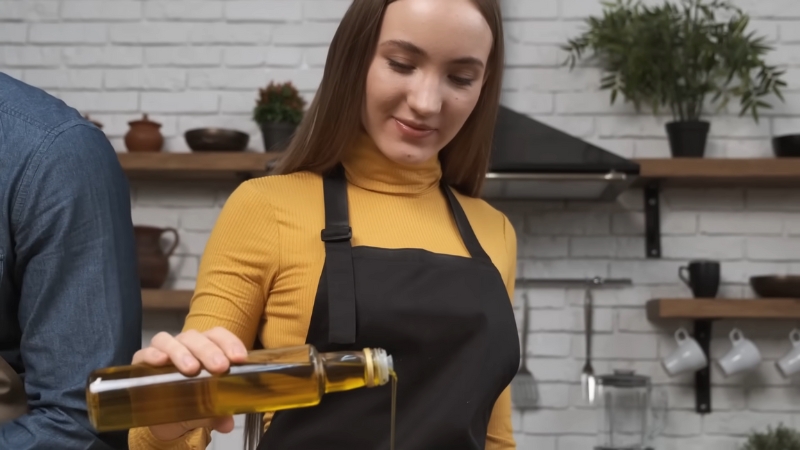 A Woman in A Yellow Sweater and Black Apron Pours Cooking Oil from A Bottle in A Kitchen Setting