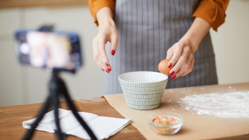A person cracking an egg into a bowl while filming cooking videos with a smartphone