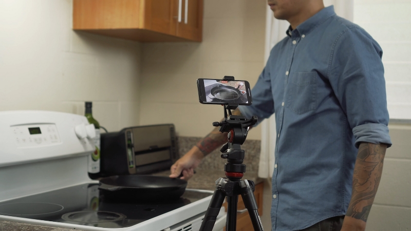 A person filming a cooking video with a smartphone on a tripod while cooking on the stove