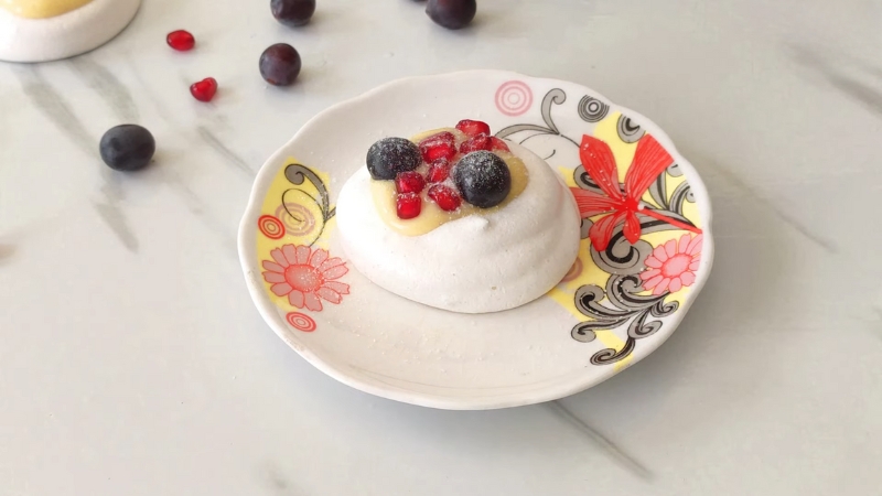 A mini pavlova topped with lemon curd, blueberries, and pomegranate seeds on a decorative floral plate