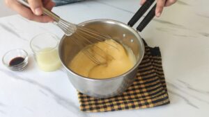 A person is whisking lemon curd in a saucepan placed on a checkered towel, with lemon juice and vanilla extract in small containers nearby