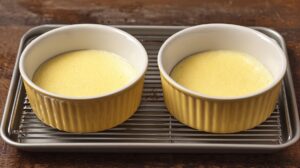 Two ramekins filled with custard mixture, placed on a wire rack over a baking sheet