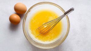 Glass bowl with whisk, egg yolks, and sugar being mixed, with two whole eggs nearby