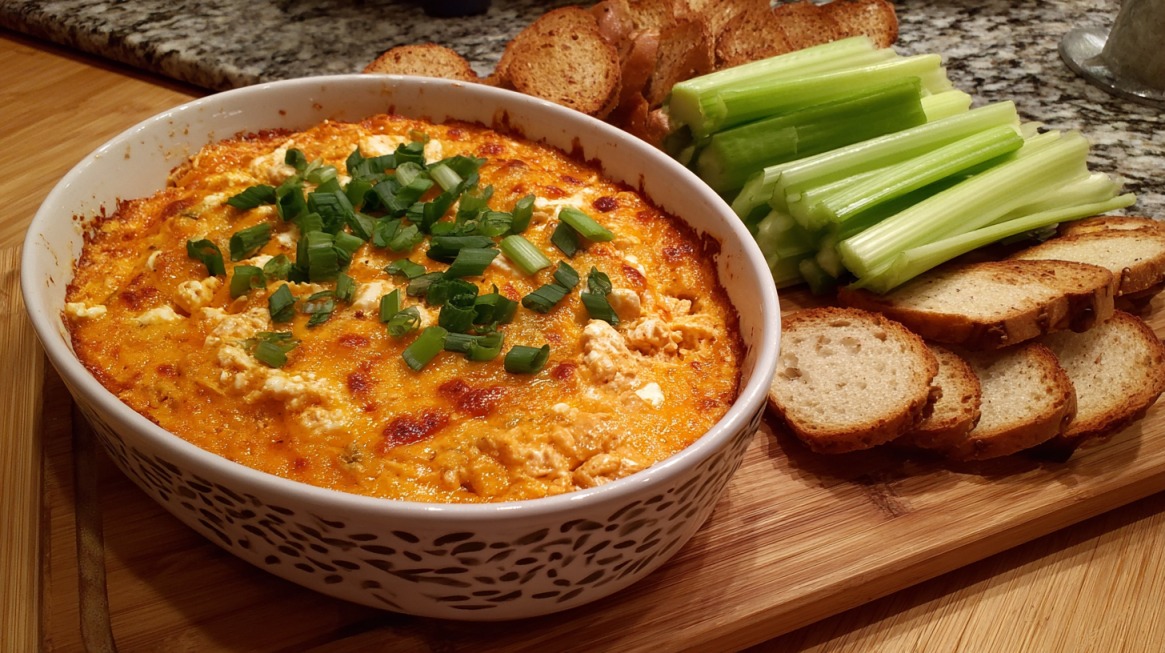 A baking dish filled with golden-brown buffalo chicken dip topped with chopped green onions, served on a wooden board with sliced bread and celery sticks