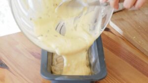 Cake batter is being poured from a glass bowl into a greased loaf pan