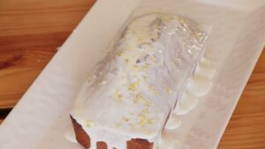 Lemon Lavender Loaf Cake cooling on a white platter with lemon glaze dripping down the sides and lemon zest sprinkled on top