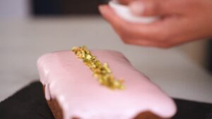 A hand sprinkles chopped pistachios onto a loaf cake covered in pink glaze