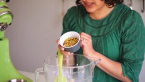 A woman holding a small bowl of chopped pistachios over a mixing bowl