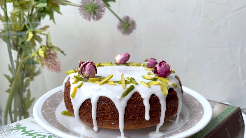 Pistachio rose cake topped with white glaze, chopped pistachios, and dried rosebuds on a plate beside a vase of flowers