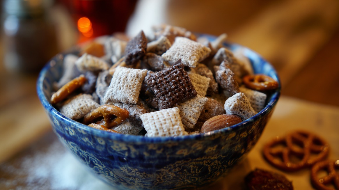 Close-up of a blue bowl filled with Puppy Chow snack mix made of cereal squares coated in powdered sugar, chocolate, and peanut butter, with pretzels and nuts mixed in