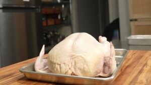 A raw whole chicken sits on a metal baking tray on a wooden countertop
