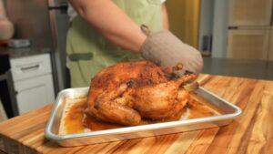 A person sets down a roasted whole chicken in a baking tray to rest on a wooden counter