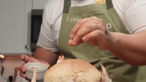 A person wearing an apron sprinkles seasoning over a whole raw chicken