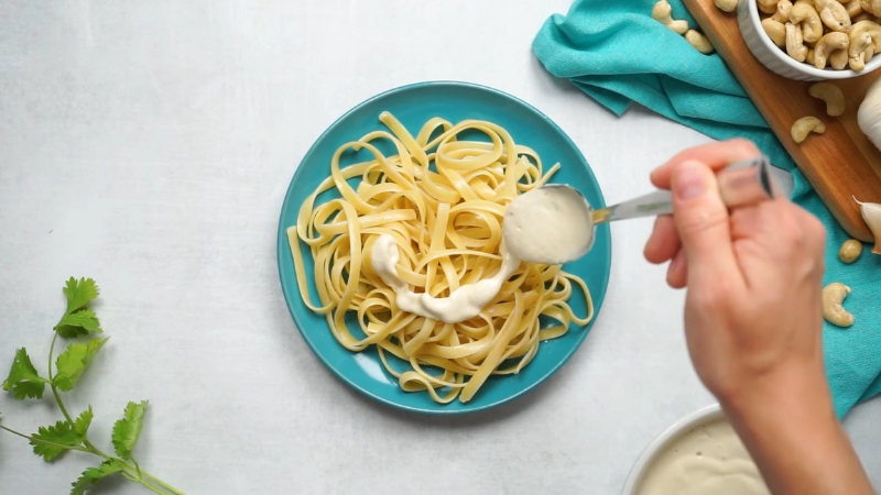 Creamy vegan cashew sauce being spooned over cooked pasta on a blue plate