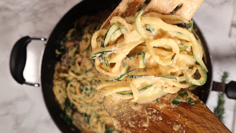 Zucchini noodles coated in a creamy cashew sauce being lifted from a pan with wooden tongs