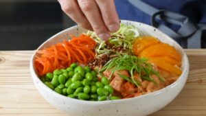 A colorful lox poke bowl topped with salmon, edamame, shredded carrots, citrus slices, and fresh garnishes being finished with a sprinkle of seasoning
