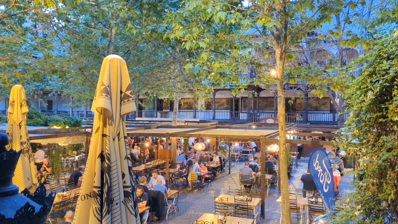 Outdoor courtyard dining at Hanu’ lui Manuc restaurant in Bucharest with guests seated under trees and umbrellas