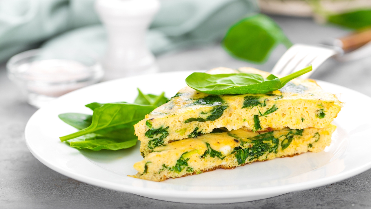 Spinach omelet slices are served on a white plate beside fresh spinach leaves