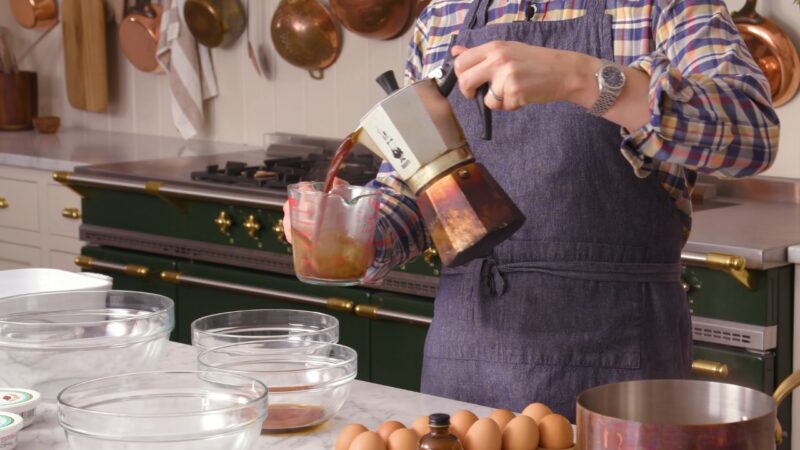 Baker pours coffee out of a moka pot into a glass cup beside bowls and eggs